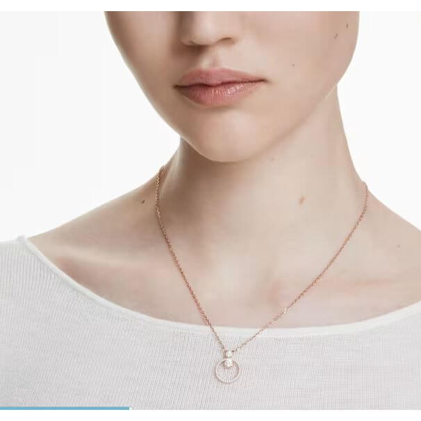 Woman wearing Swarovski necklace with circular pendant on gold chain, minimalist white top enhancing jewelry's elegance and style